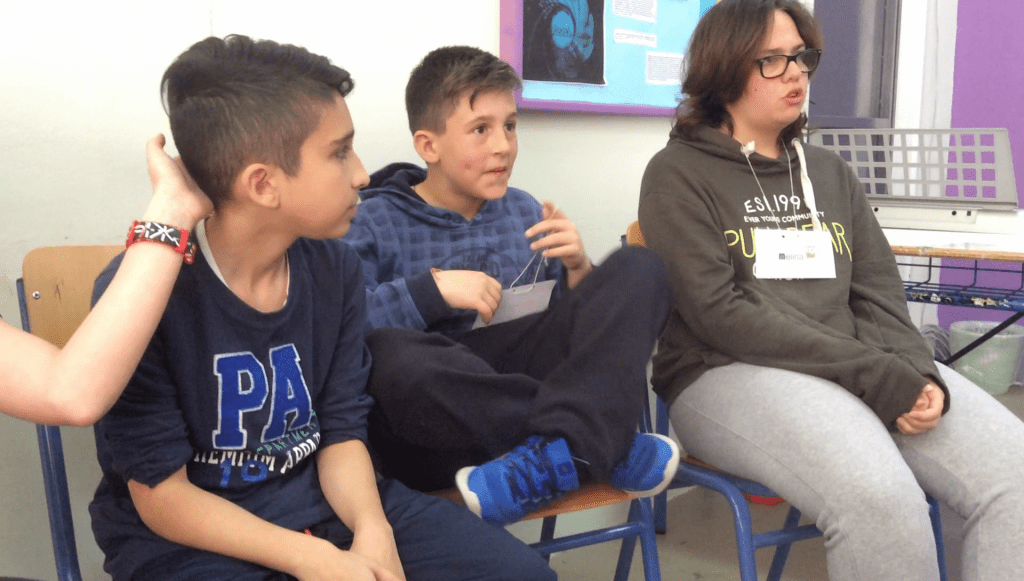Students in Greece sitting on chairs against a wall. one student is affectionately touching the head of another.