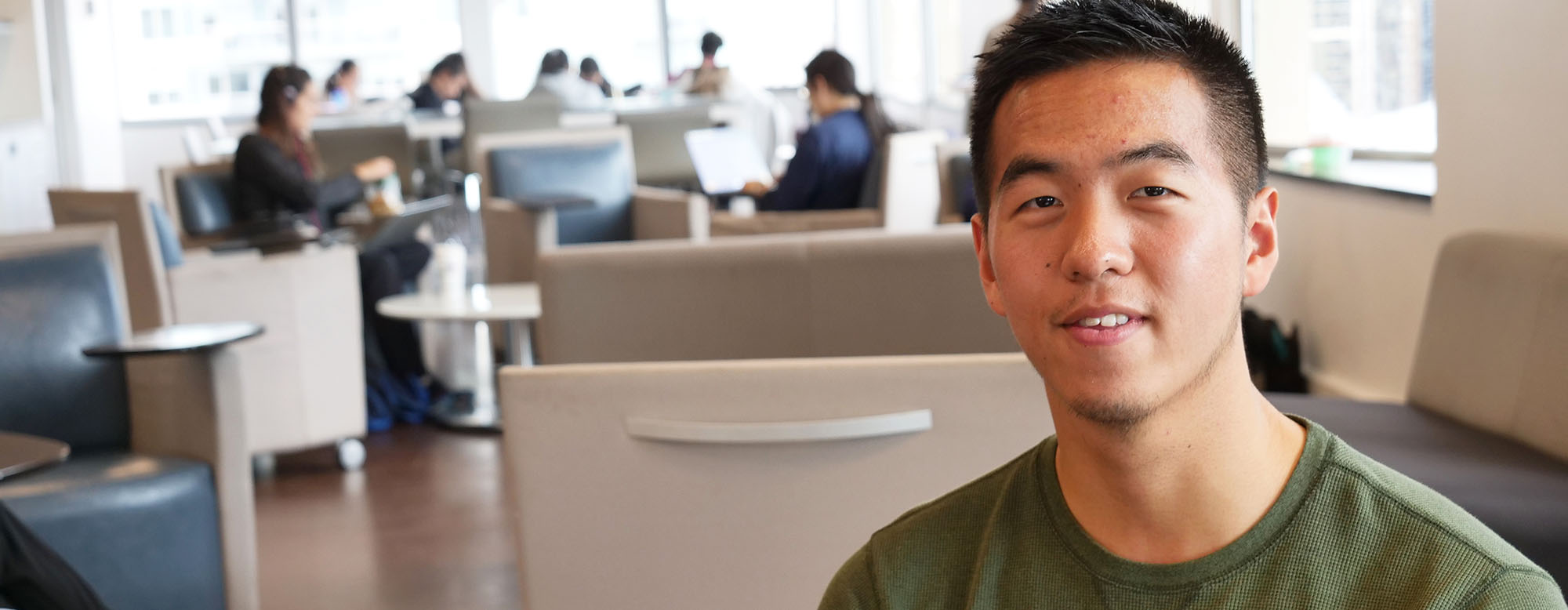 Student smiling and sitting in a room at OISE. People are working in the background.