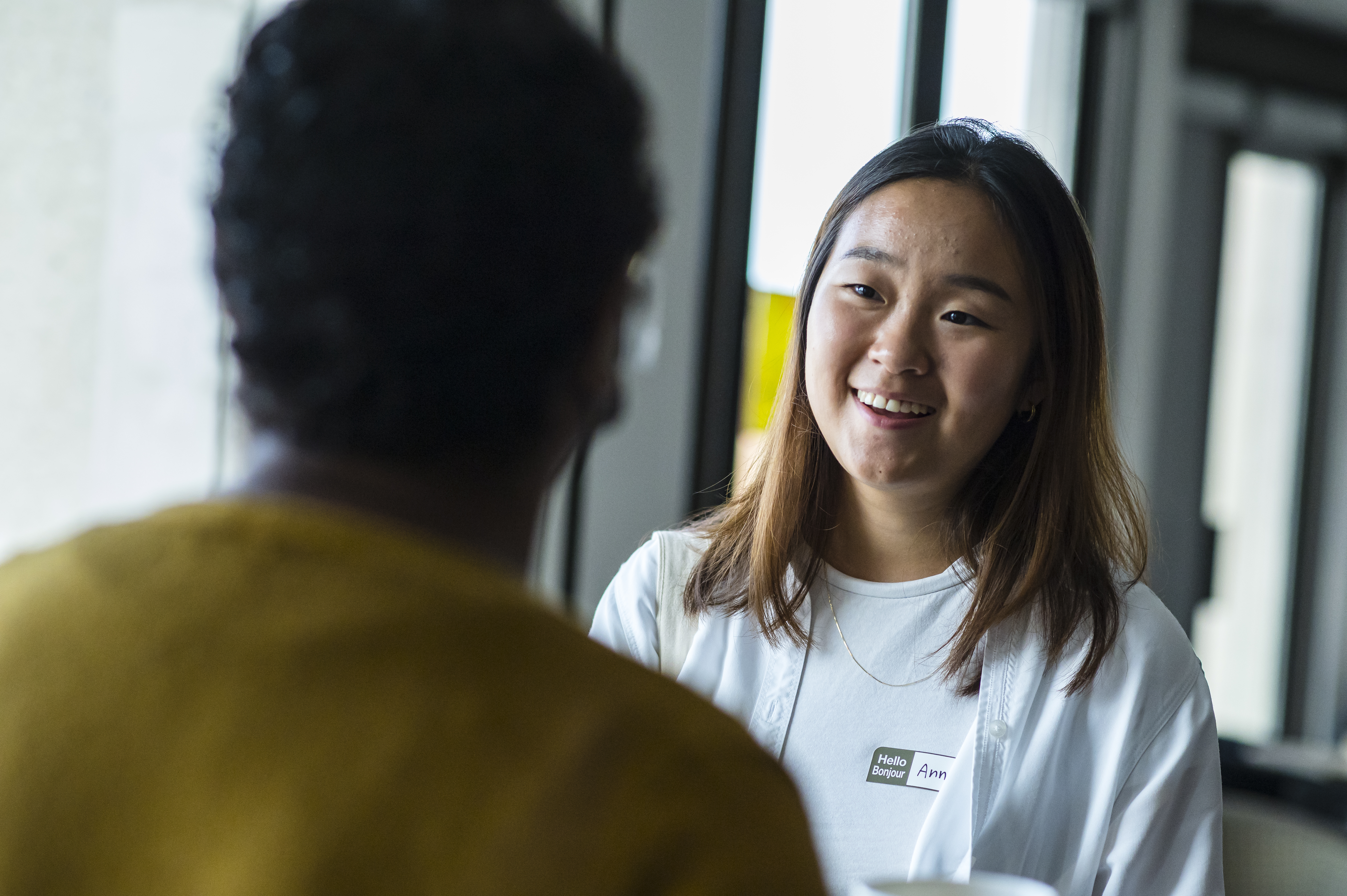 Asian woman smiles as she speaks to someone in a yellow shirt, with their back to the camera
