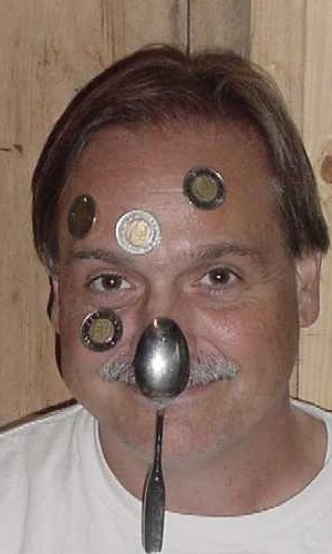 Image of a smiling man with a spoon hanging from his nose, and coins stuck to his face