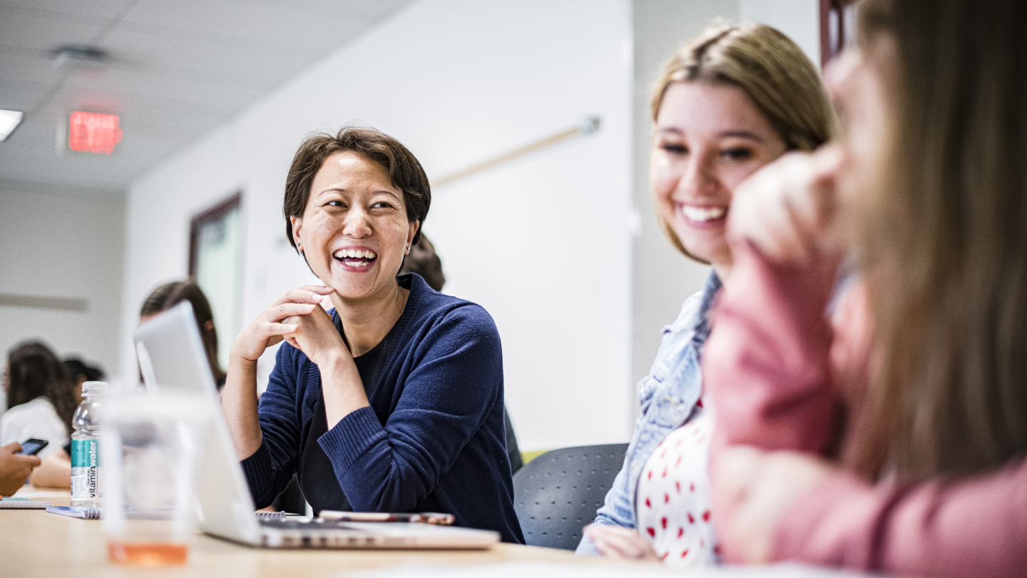 Two women, one in a dark blue sweater, the other in a denim jacket, are laughing in a classroom. One has their laptop open in front of them. A third woman in a pink sweater can be seen out of focus.