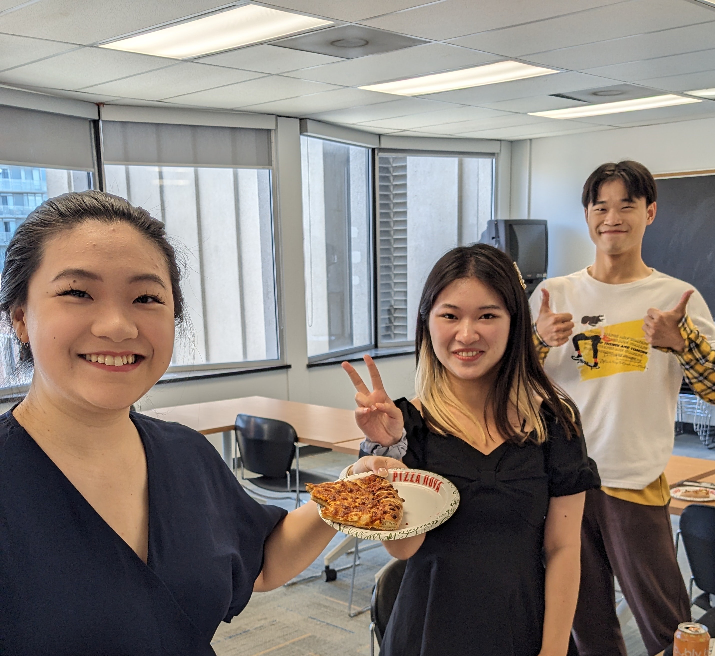 Three people are smiling, one holds pizza, one is giving two thumbs up, and one is making a peace sign.
