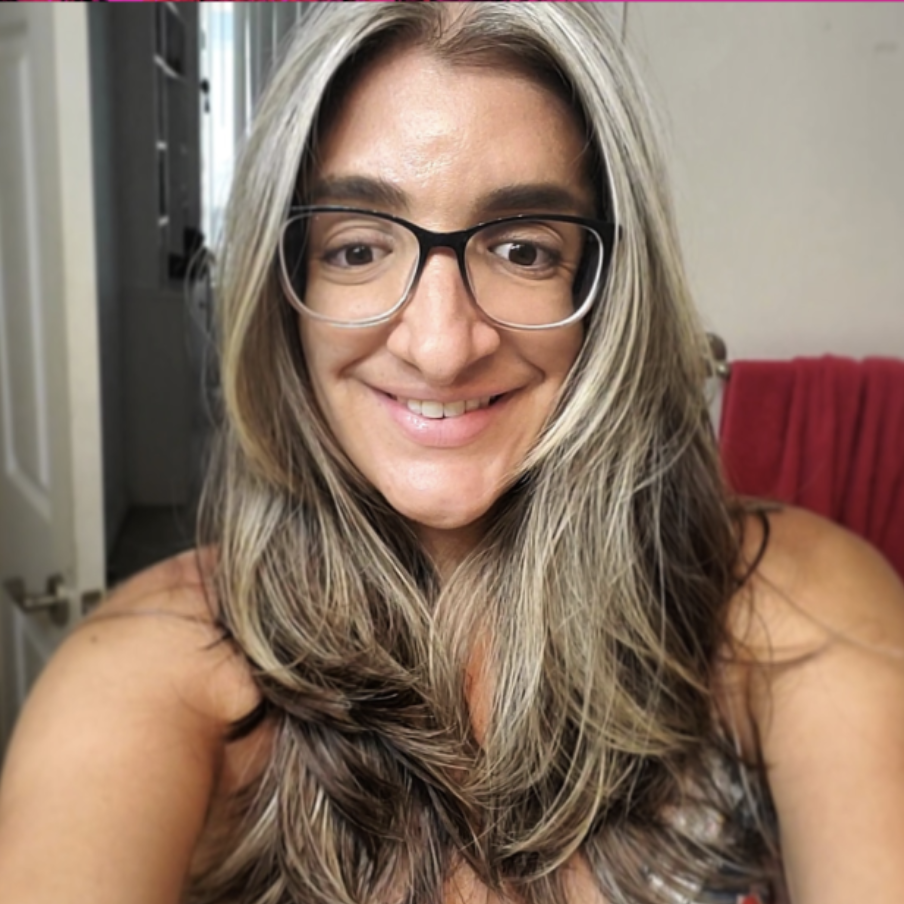 Tania Ruiz-Chapman is smiling with long grey and black hair and black glasses. Her hair covers her top, which appears to be floral. A red towel hangs in the background and a door is ajar. 