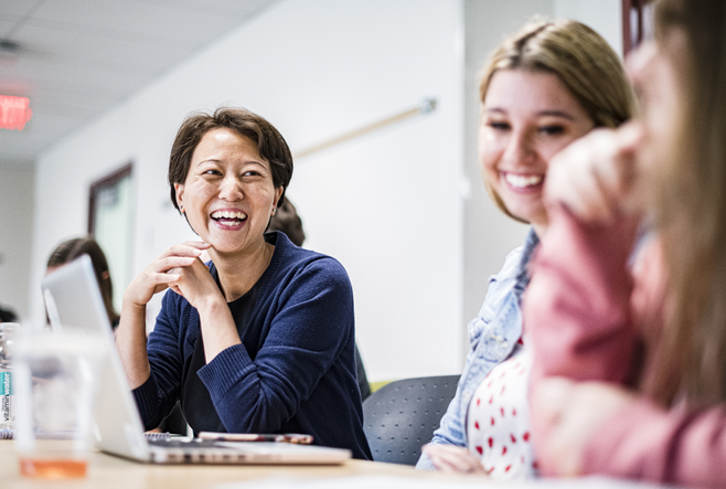 Two women, one in a dark blue sweater, the other in a denim jacket, are laughing in a classroom. One has their laptop open in front of them. A third woman in a pink sweater can be seen out of focus.