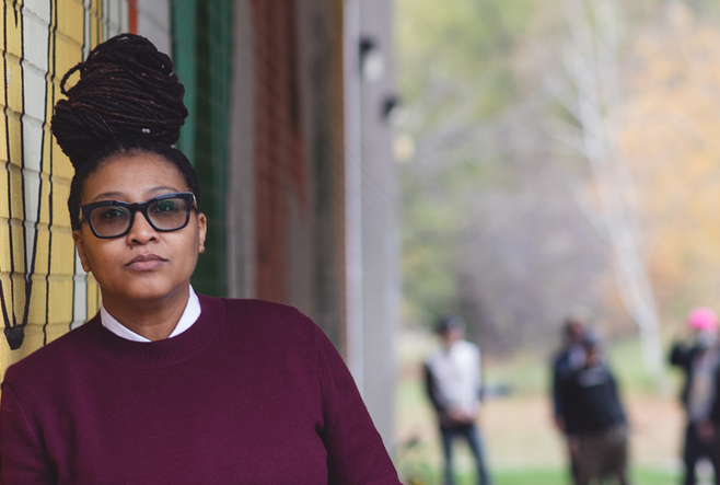 leans against a colorful mural, wearing glasses and a maroon sweater, with an outdoor background.