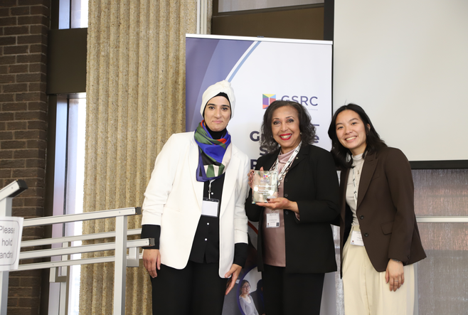 Three women standing together and smiling at the OISE Graduate Student Research Conference. The woman in the center is holding a glass award. Behind them is a banner displaying the conference logo and theme. They are dressed in professional attire and standing on a stage or presentation area."
