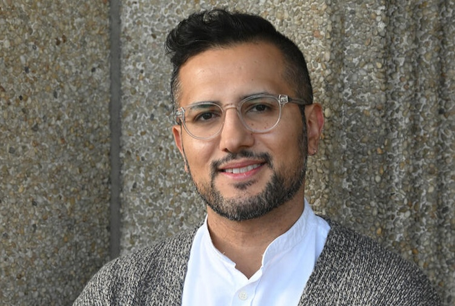 Headshot of Dr Shozab Raza standing in front of a concrete wall. He is smiling, with clear glasses and a beard, wearing a white shirt and gray cardigan.