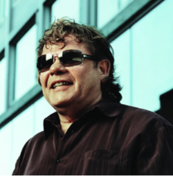 Rod Michalko wears sunglasses and a dark shirt smiles. He is in front of a glass building, looking above the camera. The photo has a blue/green hue. 