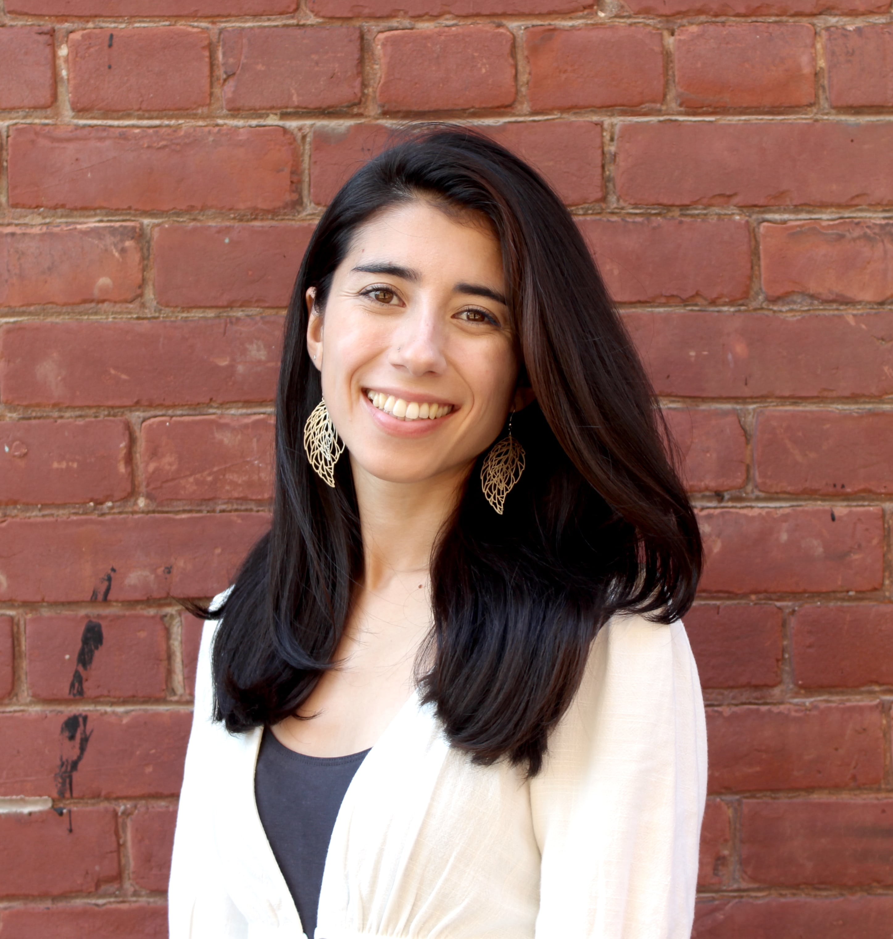 Emiko Newman stands before a brick wall, smiling. She has long straight brown hair. She wears a white blouse and large earrings. 