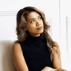 Elaine Cagulada sits on a beige seat before a white background and has a small smile, looking away from the camera. She has with wavy dark blonde hair and wears a black sleeveless turtleneck. 