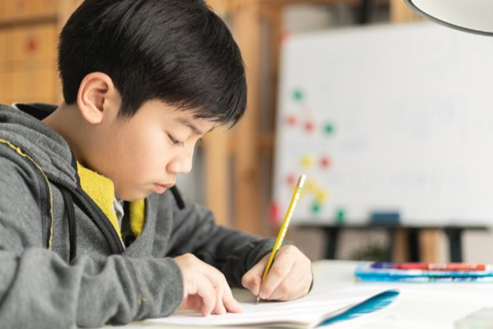 East Asian teenager is writing on a piece of paper with a pencil and looking thoughtful 