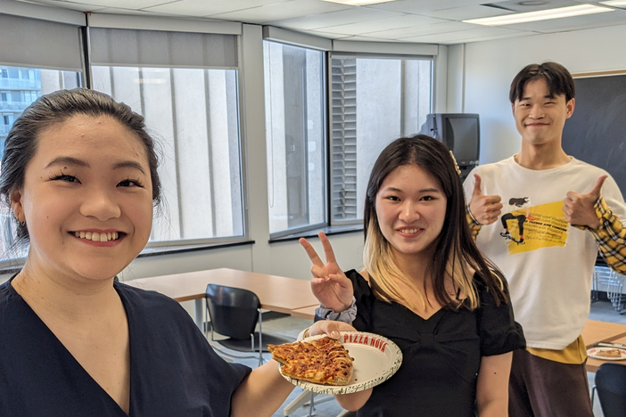 Three people are smiling, one holds pizza, one is giving two thumbs up, and one is making a peace sign.