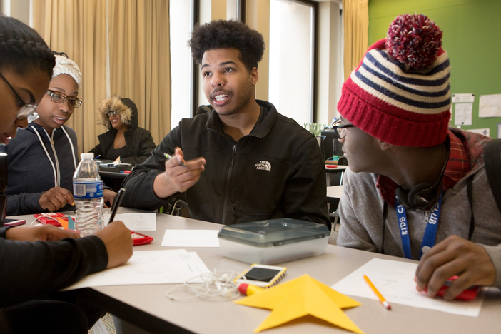 A group of three Black youth are sitting around a table in classroom setting and engaged in converation