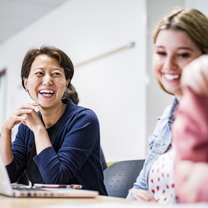 Two women, one in a dark blue sweater, the other in a denim jacket, are laughing in a classroom. One has their laptop open in front of them. A third woman in a pink sweater can be seen out of focus.