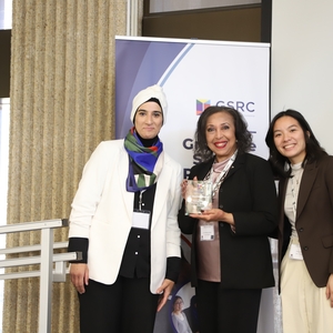 Three women standing together and smiling at the OISE Graduate Student Research Conference. The woman in the center is holding a glass award. Behind them is a banner displaying the conference logo and theme. They are dressed in professional attire and standing on a stage or presentation area."