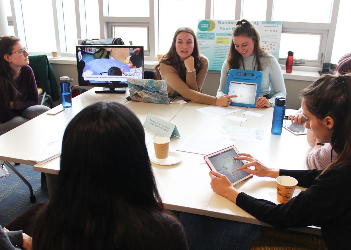 A group of people sitting around a table in a meeting room, some holding tablets and notebooks.