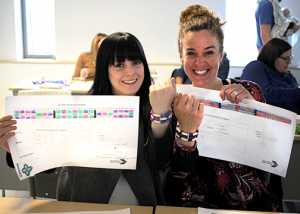 The image shows two people sitting at a table, holding up pieces of paper with colorful charts and text on them.