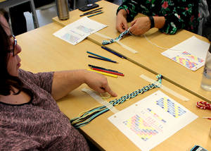 Two people sitting at a table working on a craft project involving colorful threads.