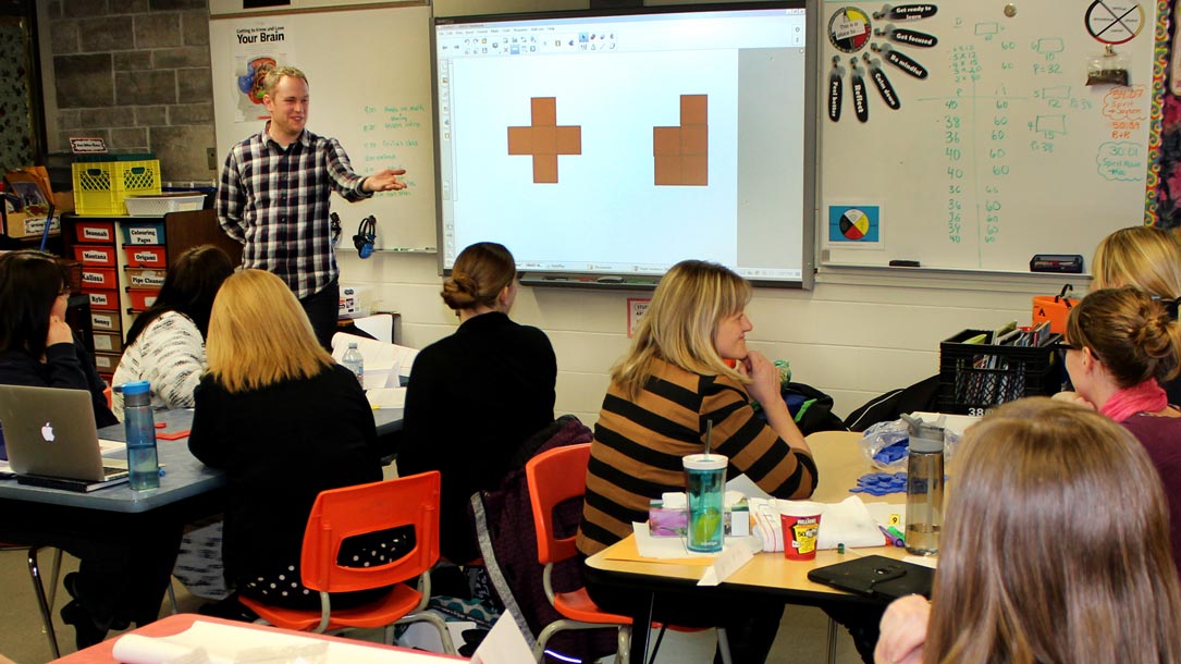 A classroom setting with students seated at tables and a teacher standing in front of a whiteboard.