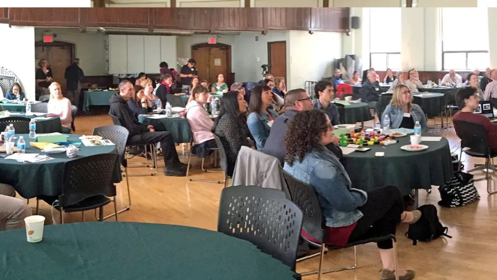 People seated at round tables in a large room, attentively listening to a speaker at a podium.