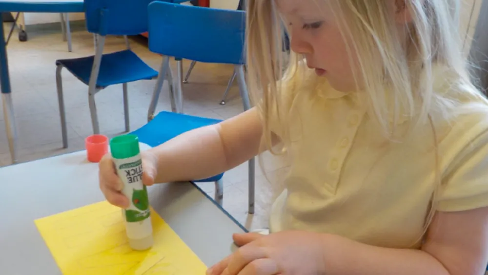 Young child using a glue stick on a yellow piece of paper at a classroom table.