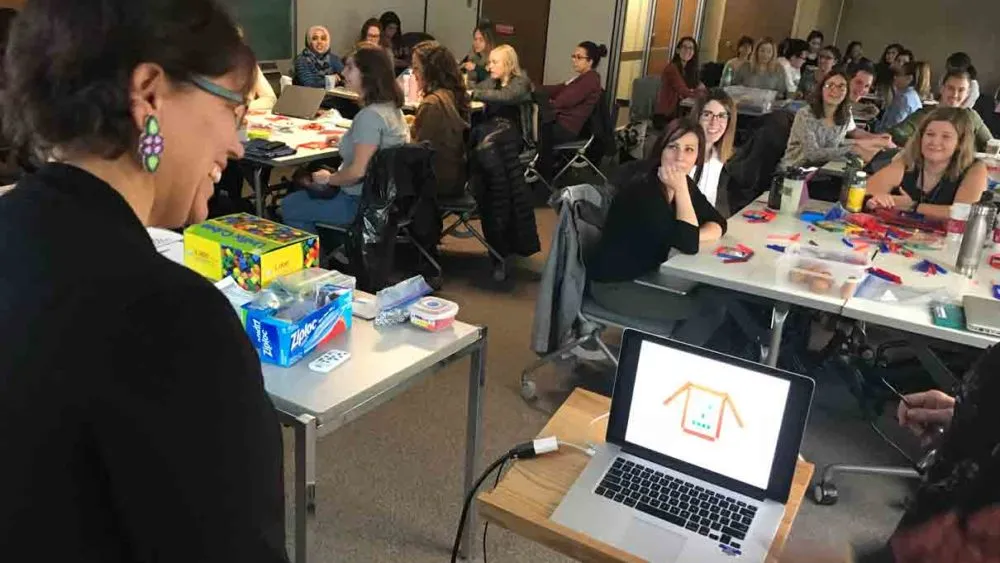 A woman presents to a room full of people seated at tables with craft supplies and laptops.