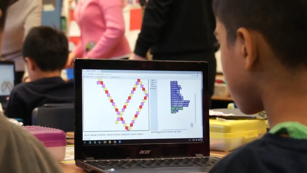 A child looks at a laptop screen displaying a colorful block-based coding project in a classroom.