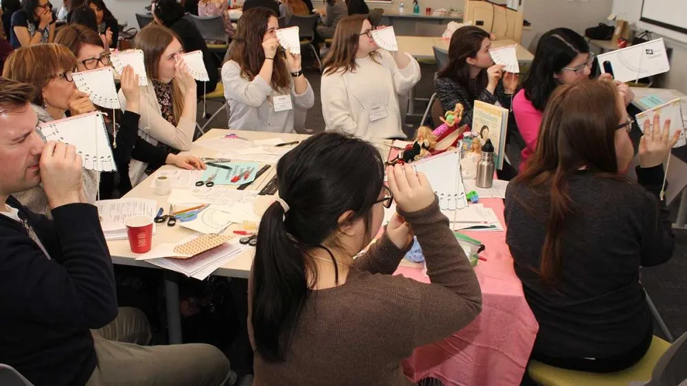 A group of people sitting around tables in a classroom or workshop setting, holding up pieces of paper with holes punched in them. The tables are covered with various supplies such as scissors, markers, and papers.