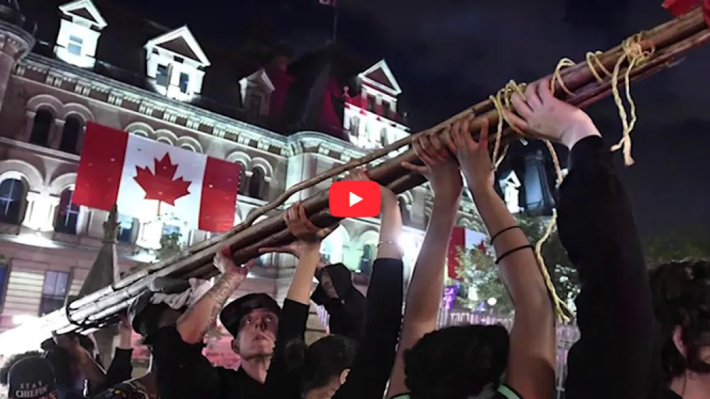 Protesters raise a large wooden pole in front of a Canadian government building at night.