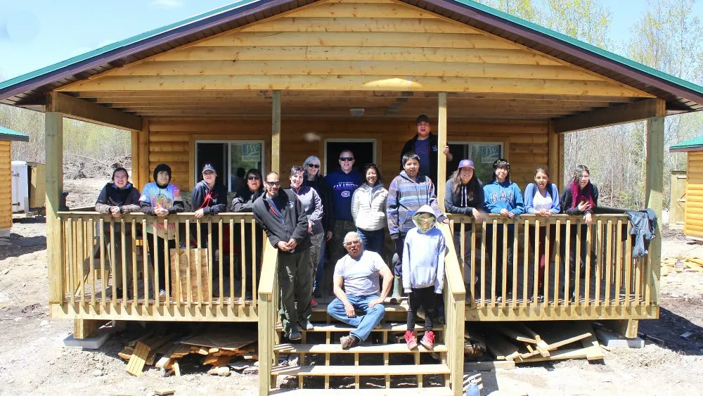 A group of people standing and sitting on the porch of a wooden cabin with a green roof, surrounded by trees.