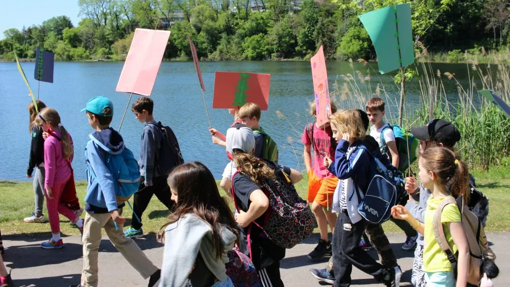 A group of children walking outdoors near a body of water, holding various colored signs.