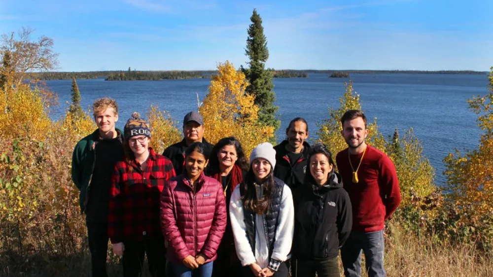 A group of nine people standing outdoors near a body of water with trees and foliage in the background.