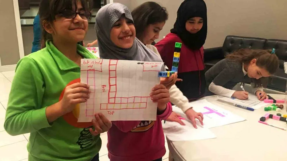 Two children holding a piece of paper with a drawing of a 3D structure made from cubes. One child is holding a stack of colored cubes. Other children are engaged in similar activities in the background.