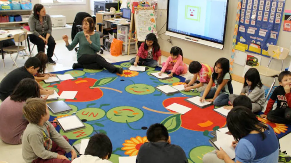 Children sitting in a circle on a colorful classroom rug, writing on clipboards while a teacher speaks.