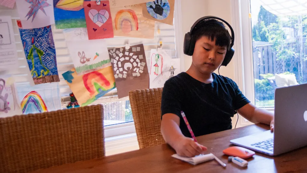 A person sitting at a wooden table, wearing headphones, and using a laptop. The person is writing on a notepad. The background features colorful drawings and artwork pinned to a wall.