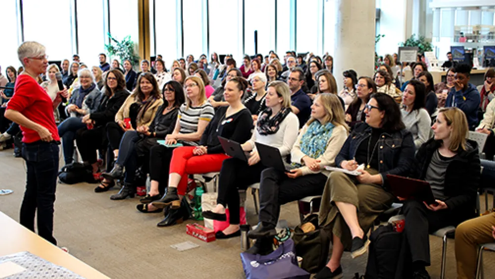 The image shows a large group of people seated in a room, attentively listening to a person standing at the front.