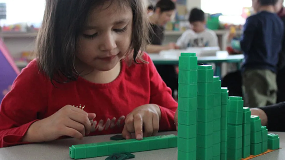 A child in a red shirt sitting at a table and playing with green building blocks. Other children are engaged in activities in the background.