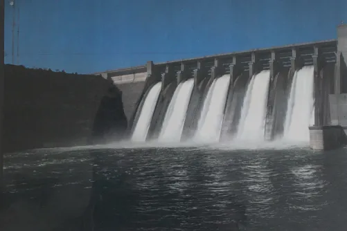 A large dam with a strong flow of water spilling over its top, set against a backdrop of greenery and blue sky.