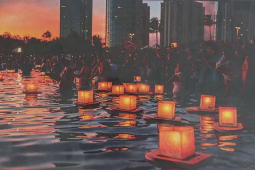 Lanterns float on water at sunset, with a crowd gathered. Tall buildings are visible in the background, silhouetted against the colorful sky.