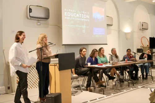 A panel of speakers seated at a long table with two women standing at a podium, presenting on global climate education. A screen behind them displays the presentation title.