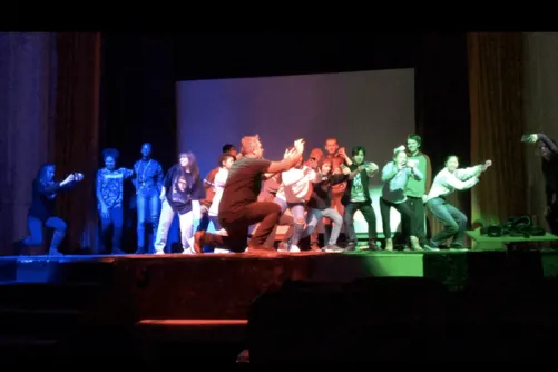 A group of people pose playfully on a stage lit by red, blue, and green lights, with one person in front holding a phone