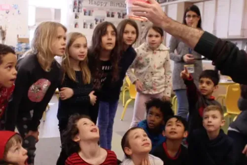 Raadiyah holding a container of salmon eggs up in the air, as a group of students stare at it Grade 2 students see the salmon eggs for the first time