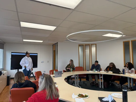 A person standing speaking in front of a group of people seated at a round table.