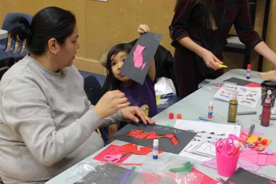 A child shows a paper art project to an adult at a craft table.