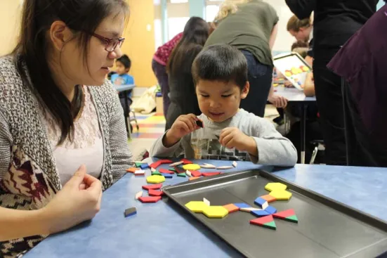 A woman watches a young boy play with colorful pattern blocks at a table.