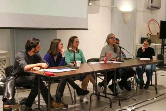 Six people sit at a long table in a panel discussion, with one woman speaking into a microphone.