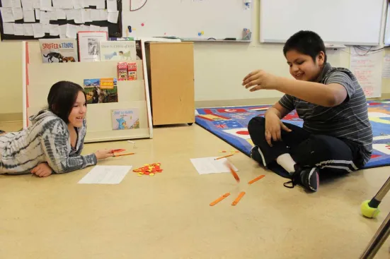 Two children play a game with orange sticks and red and yellow chips on a classroom floor.