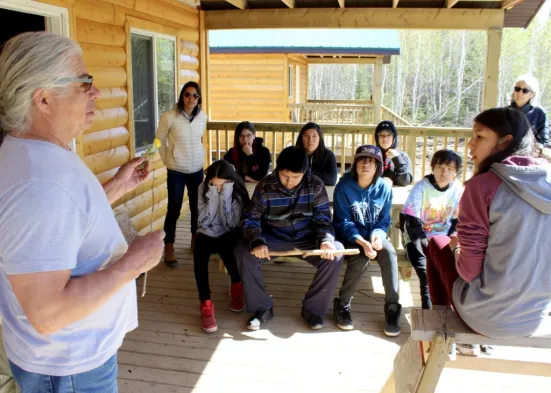 A group of people sitting on the porch of a wooden cabin. One person is standing and speaking to the group. Trees are visible in the background.