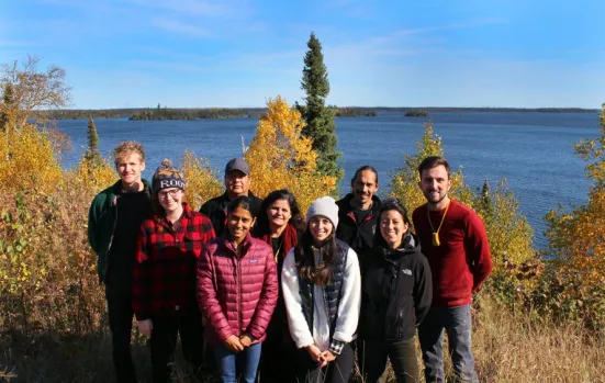 A group of nine people standing outdoors near a body of water with trees and foliage in the background.