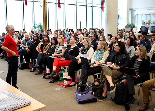 The image shows a large group of people seated in a room, attentively listening to a person standing at the front.